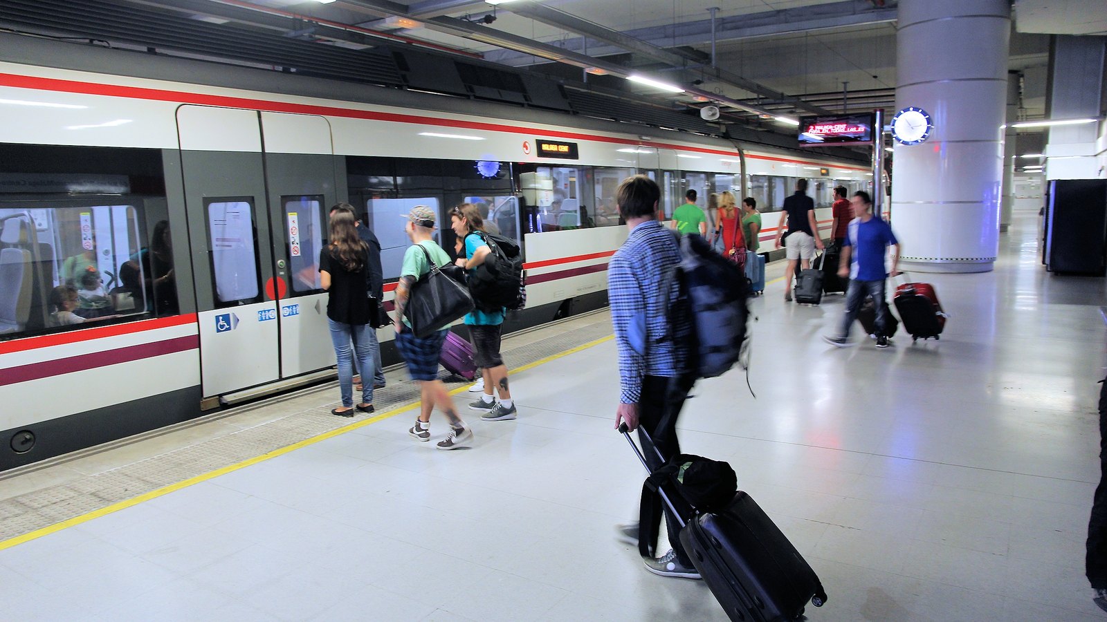 Airport passengers carrying and rolling luggage beside a train at Málaga Airport station