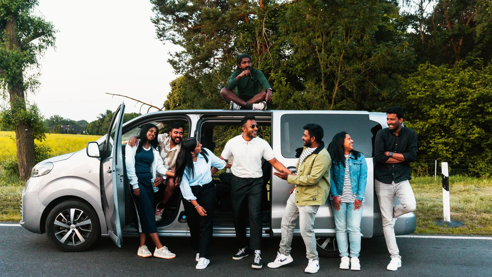 Eight adult friends smiling beside a passenger van during a relaxed road trip