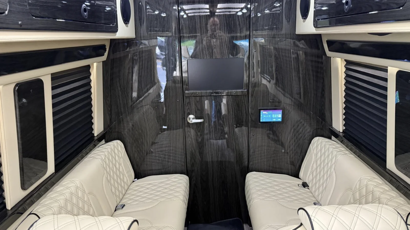 Rear cabin of the listed 2025/26 Midwest Automotive Designs van: cream quilted seats facing a glossy partition with a screen and control panel.