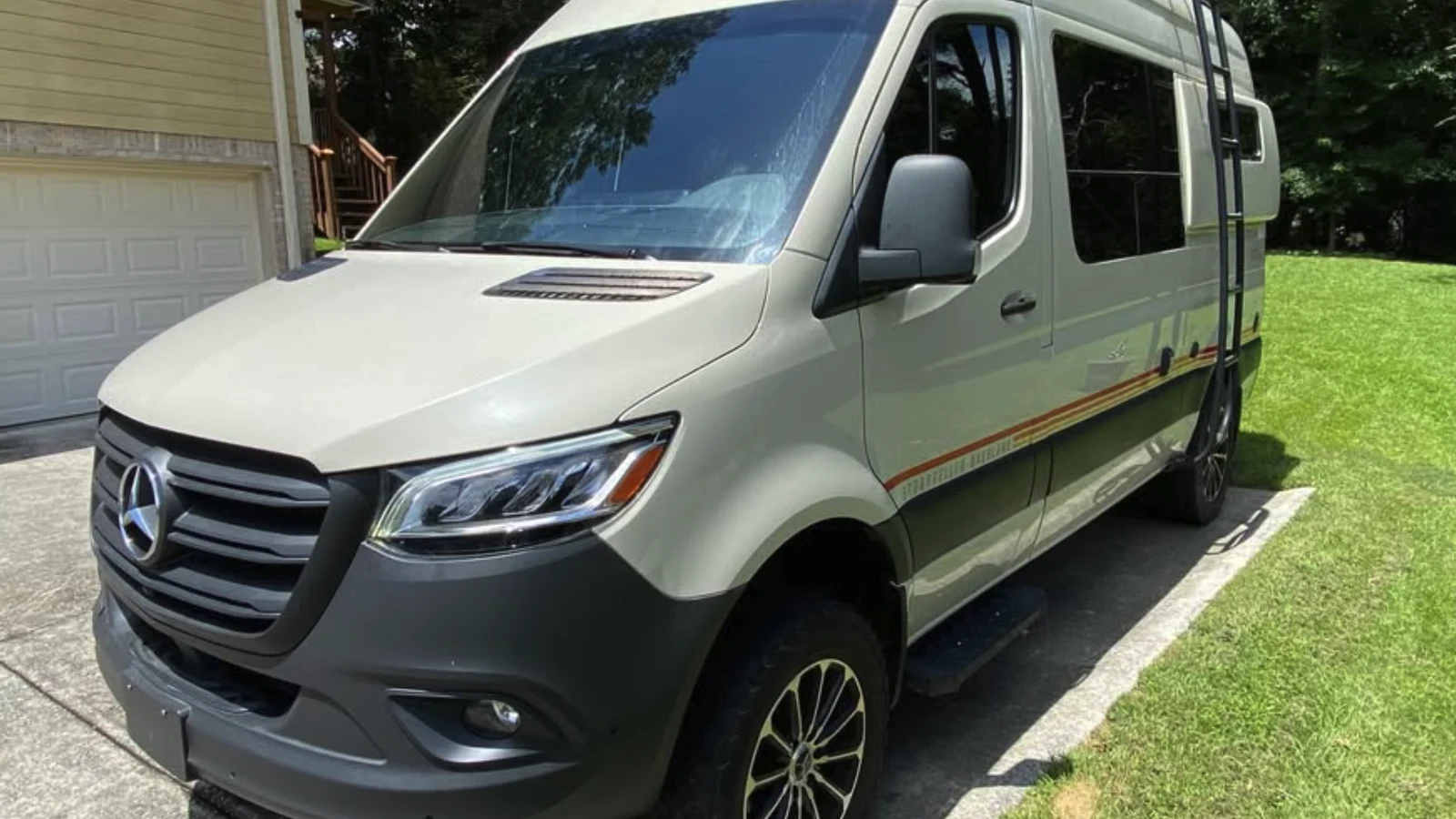 Front three-quarter view of the listed 2022 Storyteller MODE 4x4 Sprinter in a light sand color with orange side stripes and a side ladder, parked on a driveway beside a lawn.